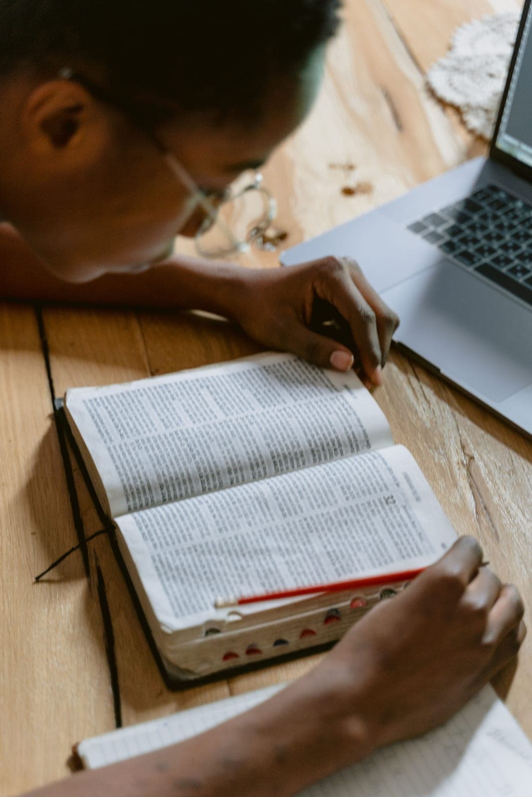 A person turning the pages of a tabbed study Bible beside an open notebook and laptop on a wooden desk