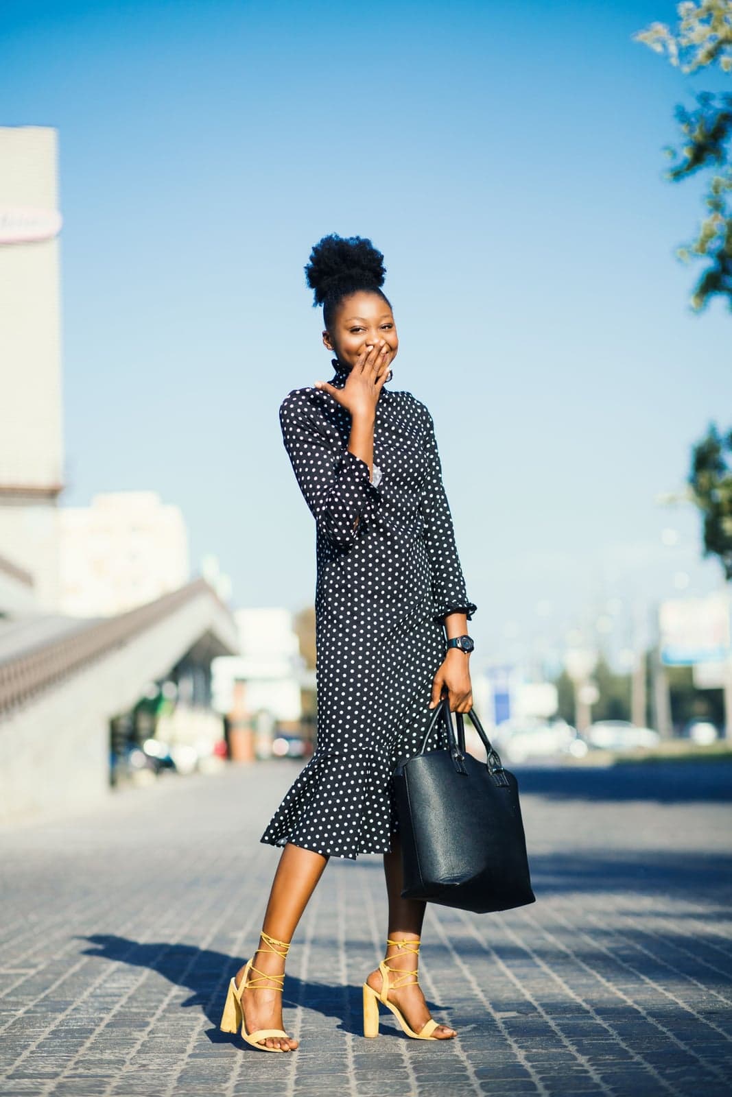 A woman in a black polka-dot dress laughing behind her hand, carrying a black tote and wearing yellow lace-up heels on a city street
