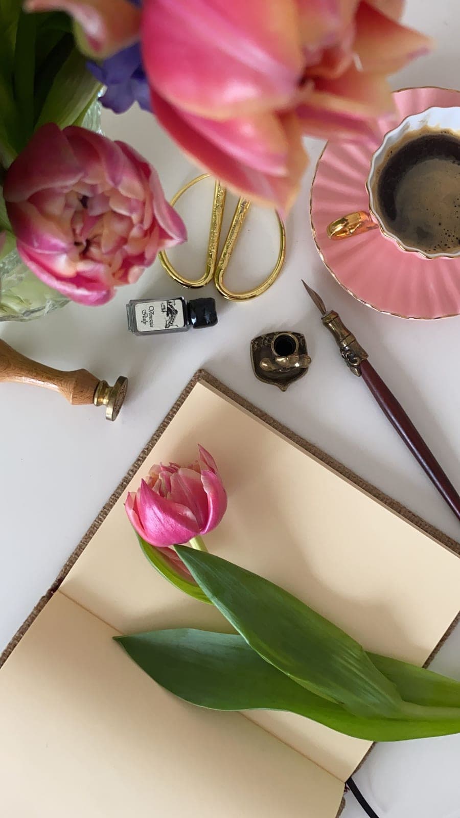 A pink tulip resting on an open cream journal beside a dip pen, ink, and a pink cup of coffee
