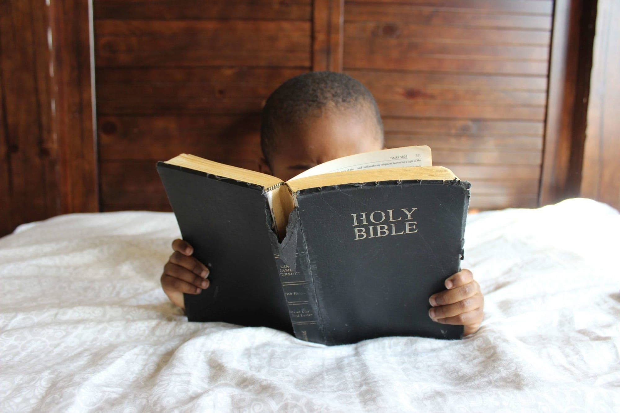 A young child sitting on a bed, absorbed in a well-worn Holy Bible held open in both hands