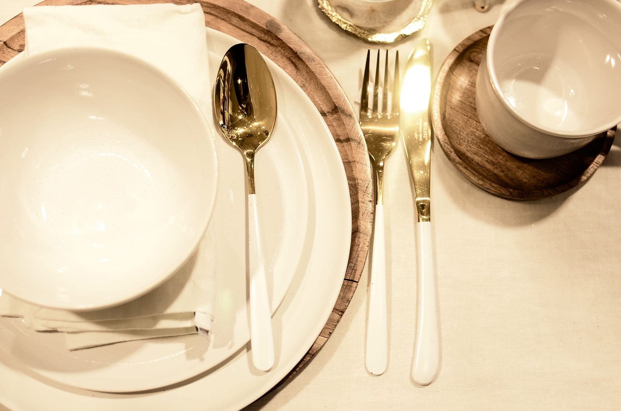 An elegant place setting: white plates on a wooden charger, gold cutlery, a linen napkin, and stoneware cups