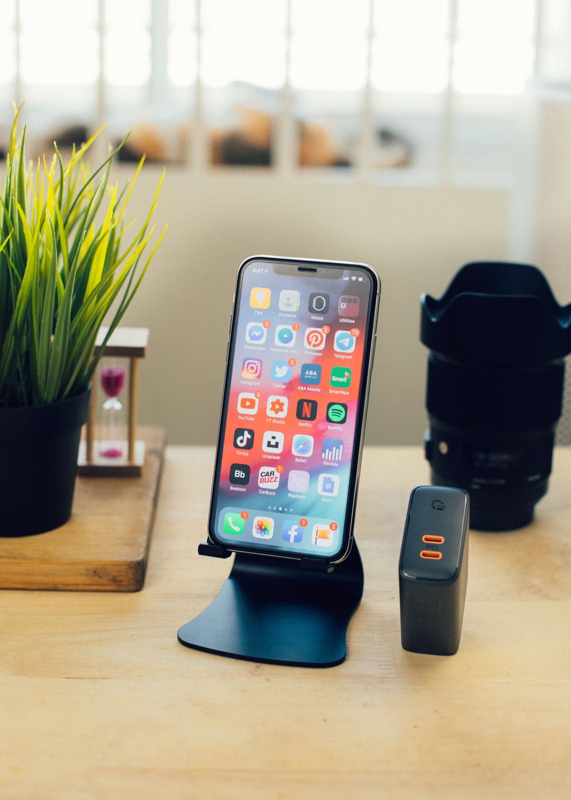 An iPhone on a desk stand showing a home screen full of messaging and social apps, beside a small plant and a camera lens