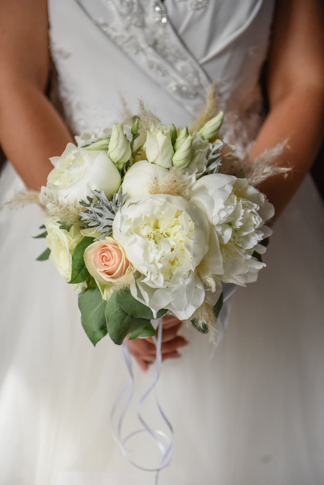 A bride holding a bouquet of white peonies, roses, and pampas grass