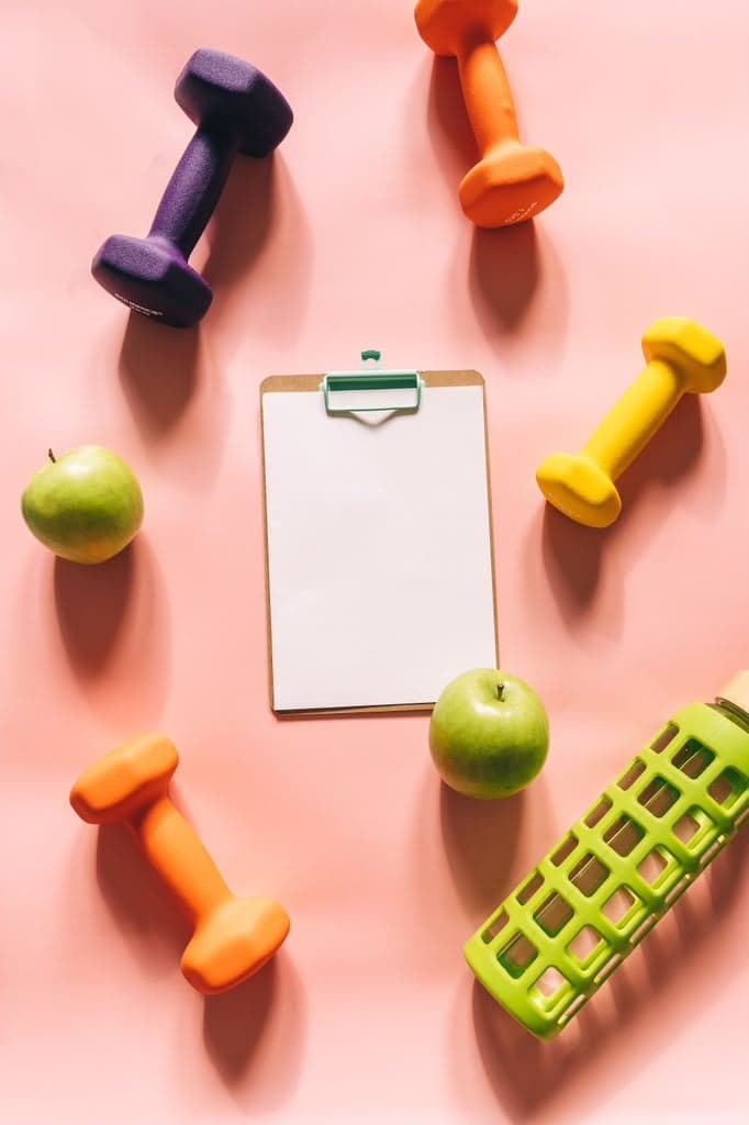 Colorful dumbbells, green apples, and a blank clipboard arranged on a pink background